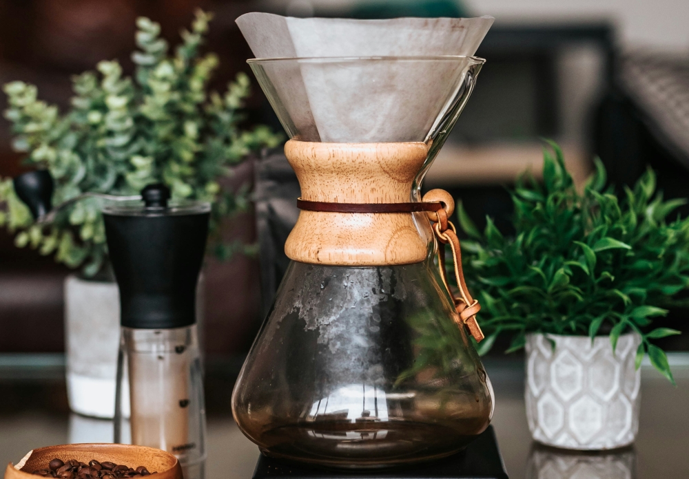 A carafe with coffee and a chemex sits on a table next to a hand ginder and some small potted plants