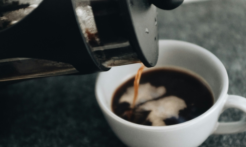 Coffee pouring out of a black french press into a small coffee cup.